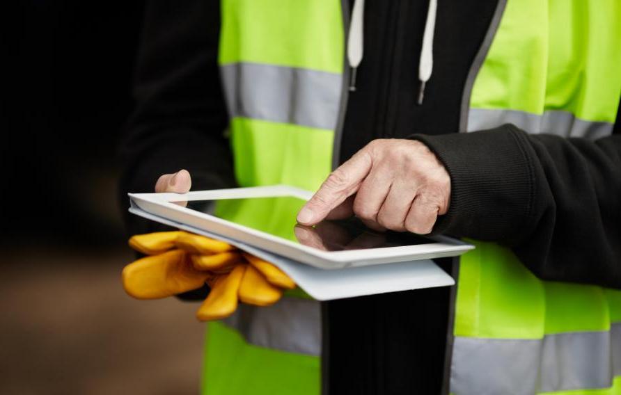 Man in yellow PPE vest holds yellow work gloves and uses an iPad